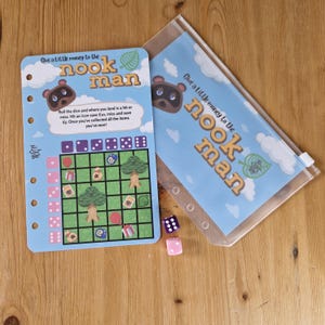 May include: A blue and white board game with the text "Give a Little money to the nook man". The game board has a grid with icons and a dice roll area. There are also two dice, one pink and one purple, on a wooden surface.
