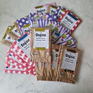 May include: A selection of "Oopsie" mystery saving challenge packs. The packs display diverse patterns, such as polka dots and stripes, alongside festive graphics. The text on the packs states "Mystery Saving Challenge 5-Pack."