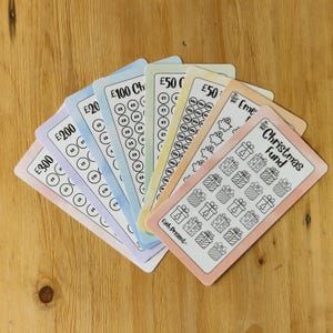 May include: A set of colourful printable Christmas saving trackers. The trackers are designed to help you save money for Christmas gifts. Each tracker has a different amount of money to save, from £50 to £300. The trackers are printed on white paper with a watercolour background.