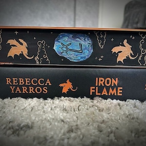 May include: Two hardback books stacked on top of each other. The top book has a black cover with a blue and silver design and two orange dragons. The bottom book has a black cover with the title "Iron Flame" and the author's name "Rebecca Yarros" in orange lettering.