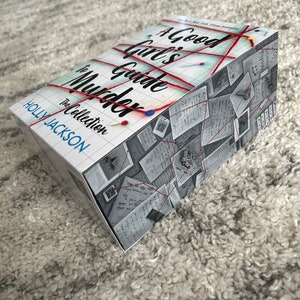 以下が含まれることがあります： ホリー・ジャクソン著「A Good Girl's Guide to Murder」というタイトルの本。表紙には、手書きのメモとグリッドパターンが書かれた紙を赤い糸でつないだものが描かれている。