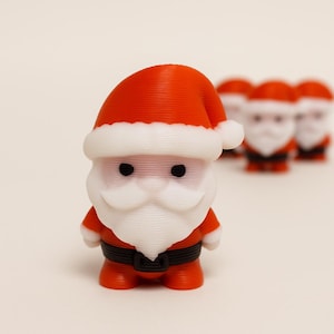 Könnte beinhalten: Eine kleine, 3D-gedruckte Weihnachtsmann-Figur mit einer leuchtend roten Mütze mit weißem Rand, einem weißen Bart, schwarzen Augen und einem schwarzen Gürtel mit Schnalle. Die Figur hat eine glatte, geschichtete Textur und drei identische Figuren dahinter.