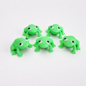 Könnte beinhalten: Fünf kleine, hellgrüne Froschfiguren, die in einer Gruppe angeordnet sind. Jeder Frosch hat eine glatte, matte Textur, vier gespreizte Beine und markante weiße Augen mit kleinen schwarzen Pupillen. Die Frösche sind in einer Gruppe platziert.