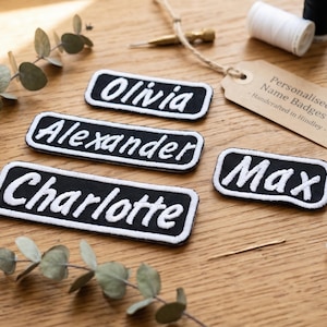 May include: Four rectangular black fabric patches with white embroidered borders and white text reading Olivia, Alexander, Charlotte, and Max. A tan paper tag with the text Personalised Name Badges Handcrafted in Hindley is attached by twine.