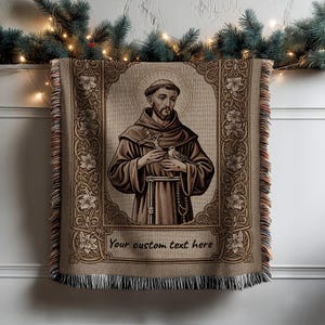 Könnte beinhalten: Eine beigefarbene Wurfadecke mit einem religiösen Bild eines Mannes in braunen Gewändern, der ein Kreuz und einen Vogel hält. Das Bild ist mit floralen Mustern umrahmt und der Text "Your custom text here" steht unten. Die Decke hat einen Fransenrand.