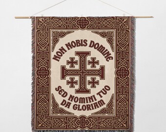 May include: Woven wall hanging with a cream background and a brown border featuring a cross design. The text "NON NOBIS DOMINE SED NOMINI TUO DA GLORIAM" is displayed in brown. The hanging is suspended from a wooden rod.