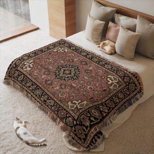Könnte beinhalten: Eine gewebte Decke mit einem detaillierten Blumenmuster in Rosa, Schwarz und Creme. Die Decke ist rechteckig mit einem Fransenrand und liegt auf einem Bett. Eine Katze schläft auf dem Bett und eine andere Katze liegt auf dem Boden.