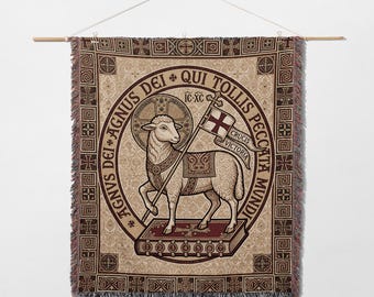 May include: A woven tapestry featuring a religious illustration of a lamb holding a flag with a cross. The lamb stands on a book within a circular design with Latin text. The tapestry has a beige background with a repeating pattern and a fringed edge.