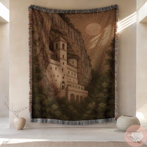 May include: A woven tapestry depicting a stone monastery built into a cliff face. The artwork is in shades of brown and beige, with a sunburst design in the upper right. The tapestry has a fringed border, suitable for wall hanging.