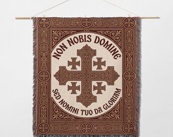May include: Woven wall hanging featuring a large cross design in shades of brown and cream, with the Latin phrase "Non Nobis Domine" and "Sed Nomini Tuo Da Gloriam" in a circular pattern. The tapestry is hung from a wooden rod.