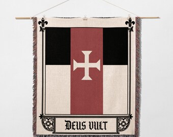 May include: Woven wall hanging featuring a design with a white cross on a red vertical stripe, flanked by black and white sections. The phrase "Deus Vult" is displayed in a decorative banner at the bottom. The edges have a fringed border.