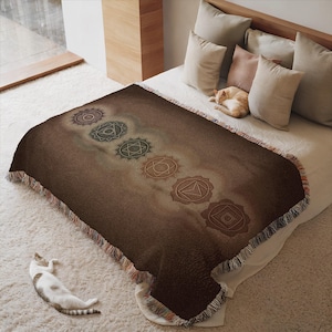 Könnte beinhalten: Braune Decke mit sieben Chakra-Symbolen. Die Decke hat einen Fransenrand und wird auf einem Bett mit Kissen präsentiert. Eine Katze liegt auf dem Bett und eine andere Katze liegt auf dem Boden.