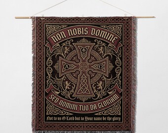 May include: A tapestry with a dark background and a large cross design. The cross is in the center, with text above and below it. The text reads "NON NOBIS DOMINE" and "SED NOMINI TUO DA GLORIAM". The tapestry is hanging from a wooden rod.