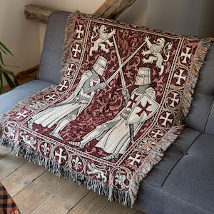 May include: A woven throw blanket featuring a deep red background with a repeating pattern of white crosses and lions. The central design depicts two knights in medieval armor engaged in a sword fight, with fringed edges along the sides.