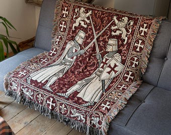 May include: A woven throw blanket featuring a deep red background with a repeating pattern of white crosses and lions. The central design depicts two knights in medieval armor engaged in a sword fight, with fringed edges along the sides.