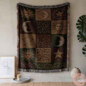May include: A woven throw blanket with a celestial design. The blanket features squares with images of the sun, moon, stars, and floral patterns in shades of brown, green, and tan. The blanket has a fringed edge.