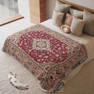 May include: A red throw blanket with a floral pattern and fringe trim. The blanket features a central medallion design and a border with repeating floral motifs. The color palette includes red, cream, black, and green. The blanket is displayed on a bed.