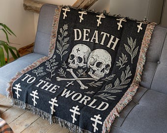 May include: A black woven throw blanket featuring two white skulls and crossbones in the center. The text "DEATH TO THE WORLD" is printed in white above and below the skulls. The design includes white botanical branches and Orthodox crosses.