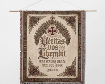 May include: A woven wall hanging with a cream background and ornate brown and burgundy detailing. The text "Veritas Vos Liberabit" is displayed in elegant script, with "The Truth Shall Set You Free" below. A wooden rod and string are used for hanging.
