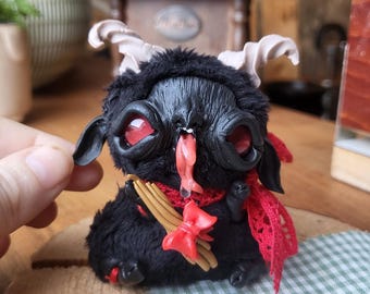 May include: A handcrafted black creature figurine with red eyes, small horns, and a red scarf. The figurine has a whimsical design, with a red bow and a small, red, tongue-like protrusion. The figure is placed on a wooden slice.