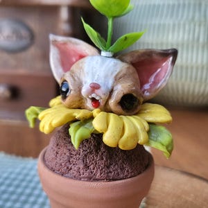 Könnte beinhalten: Eine kleine Skulptur eines Wesens mit großen Ohren und dunklen Augen, das aus einer gelben Sonnenblumenblüte hervorkommt, die auf einer braunen, strukturierten Basis in einem terrakottafarbenen Topf sitzt. Ein grüner Stängel mit Blättern und lila Blüte.