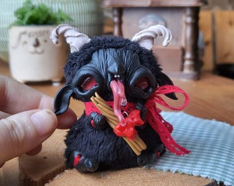 May include: A black, furry creature figurine with red eyes and a long, pink tongue. It has white horns, a red ribbon, and holds what appears to be cinnamon sticks. The figurine is displayed on a wooden slice.
