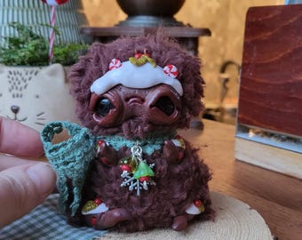 May include: A handmade brown creature figurine with large eyes, a white hat, and a green collar. It holds a small green bag and is decorated with Christmas-themed embellishments. The figurine is placed on a wooden slice.