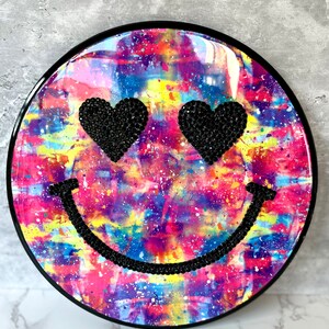 May include: A round, black-framed wall decor with a tie-dye pattern in shades of pink, purple, blue, yellow, and green. The centre of the decor features a black smiley face with black rhinestone eyes in the shape of hearts.