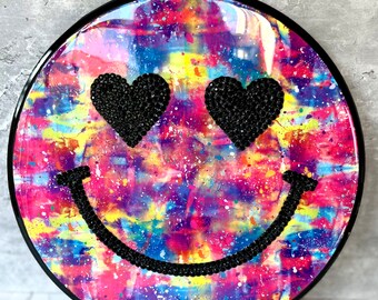 Può includere: Un decoro da parete rotondo con cornice nera e un motivo tie-dye in tonalità di rosa, viola, blu, giallo e verde. Il centro del decoro presenta un viso sorridente nero con occhi a forma di cuore in strass neri.