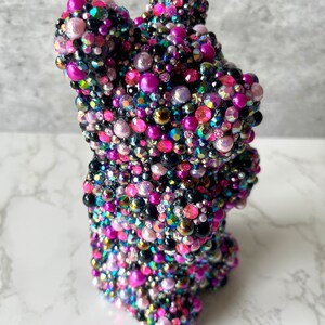 May include: A black bear figurine covered in iridescent pink, purple, and black beads. The bear is standing on a white marble surface.