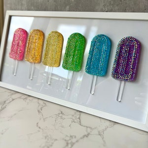 May include: A framed artwork displaying six rhinestone-covered popsicle designs. The popsicles are in pink, gold, yellow, green, blue, and purple. Each has a clear plastic stick. The artwork is presented in a white frame.