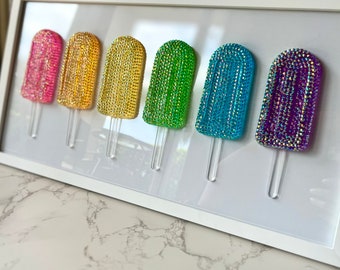 Può includere: Un'opera d'arte incorniciata con sei disegni a forma di ghiacciolo. Ogni ghiacciolo è ricoperto di strass scintillanti in diversi colori: rosa, oro, giallo, verde, blu e viola. I ghiaccioli hanno bastoncini di plastica trasparente.