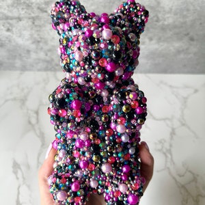 May include: A gummy bear shaped sculpture covered in black, pink, purple, and iridescent beads.
