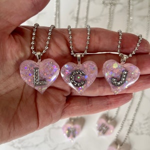 May include: Four silver chain necklaces with pink glitter heart pendants. Each heart has a different letter charm, L, C, J, and H, set with rhinestones.