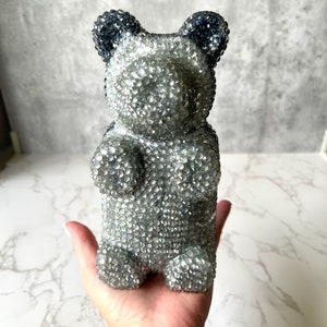 May include: A silver glitter gummy bear with black glitter ears. The bear is standing on a hand.