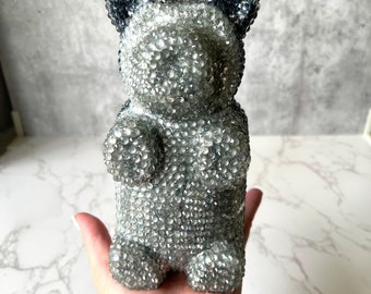 Può includere: Un orso gommoso argentato con glitter e orecchie di glitter nero. L'orso è in piedi su una mano.
