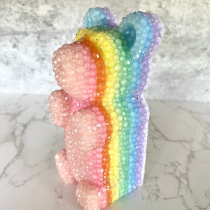 May include: A pink and rainbow glittery bear figurine. The bear is made of a clear material with a rainbow stripe down the centre. The bear is standing on a white surface.