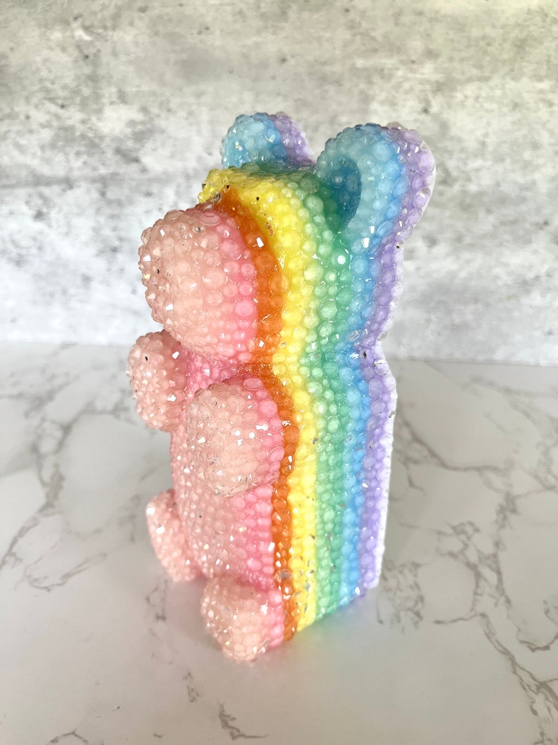 Pu&ograve; includere: Una statuetta di orso rosa e arcobaleno scintillante. L'orso &egrave; fatto di un materiale trasparente con una striscia arcobaleno al centro. L'orso &egrave; in piedi su una superficie bianca.