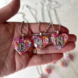 May include: Three silver necklaces with heart-shaped pendants filled with colourful sprinkles and a letter charm. The letters are D, H, and N.