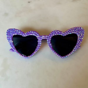 May include: Purple heart-shaped sunglasses with rhinestones. The sunglasses have black lenses.