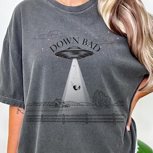 May include: Dark gray t-shirt with a graphic of a UFO beaming up a cow over a farm scene. The text "DOWN BAD" is above the UFO. The shirt is a relaxed fit.