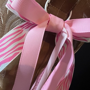 May include: A decorative bow tied to a white ring, featuring a combination of solid light pink grosgrain ribbon and a white ribbon with a bright pink zebra stripe pattern. The bow is attached to a brown suede-like material with stitched details.