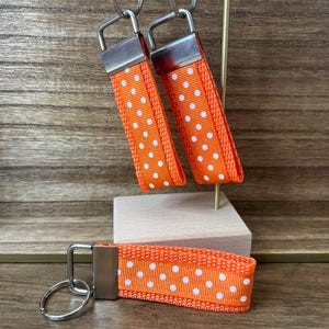 May include: Three keychains with orange fabric straps and white polka dots. Each keychain has a silver metal key ring and a silver metal clasp. The keychains are displayed on a wooden surface.