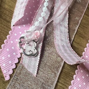 May include: Close-up of decorative ribbons in shades of pink and gold. The ribbons include a pink polka dot ribbon with a scalloped edge, a sheer pink ribbon, and a shimmering gold ribbon. A silver charm and a string of white beads are also visible.