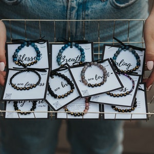 May include: A gold wire basket filled with small cards featuring beaded bracelets with positive affirmations. The bracelets are made with black lava beads and gold lettering. Some of the affirmations include "Blessed", "Breathe", "You are loved", and "You are fearless".