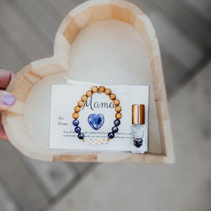 May include: A wooden heart-shaped box containing a small card, a wooden beaded bracelet with a purple heart charm, and a small bottle of essential oil. The bracelet has the word "Mama" spelled out on it.
