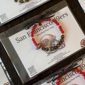 Puede incluir: Pulseras de cuentas rojas con cuentas de letras negras y doradas que deletrean nombres, y un dije de fútbol. Las pulseras están empaquetadas con tarjetas con un gráfico de casco de fútbol y el texto "San Francisco 49ers".
