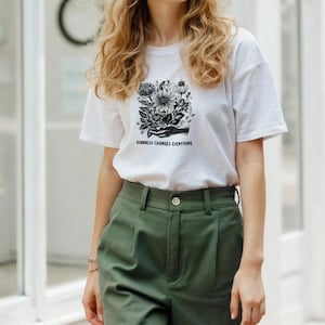 Könnte beinhalten: Weißes T-Shirt mit schwarzem Blumengrafik und dem Text "KINDNESS CHANGES EVERYTHING". Das Shirt wird mit olivgrünen Hosen getragen. Das Design zeigt eine Hand, die Blumen hält.