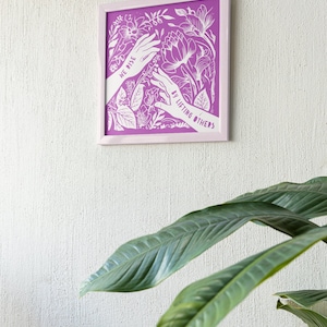 May include: A framed print with a purple background and white floral design. The print features two hands reaching towards each other with the text "We rise by lifting others".