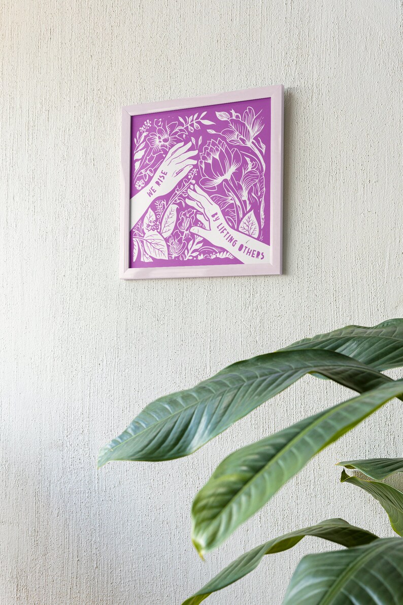 May include: A framed print with a purple background and white floral design. The print features two hands reaching towards each other with the text "We rise by lifting others".